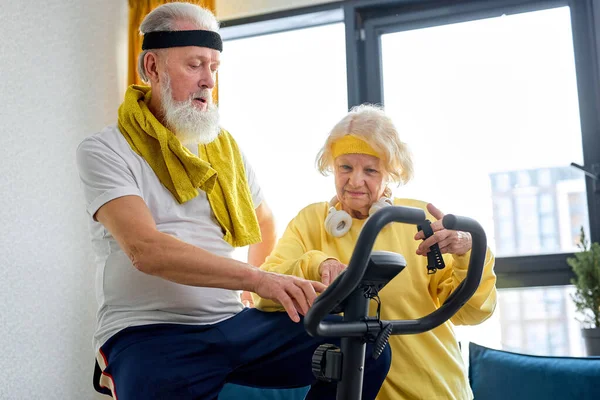woman helping man at home to do exercise on stationary bike. active couple of pensioners in sportswear help each other, gray haired male training on sport equipment in living room