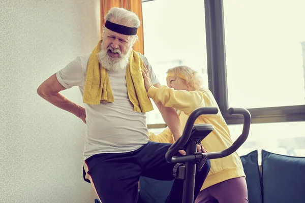 woman helping man at home to do exercise on stationary bike. active couple of pensioners in sportswear help each other, male having pain, hard to do workout on sport equipment