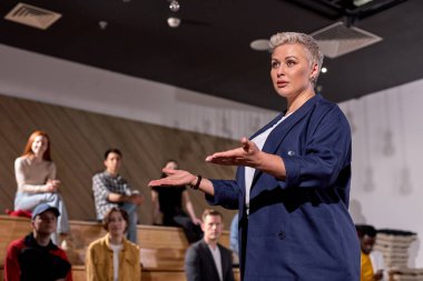 İş toplantısındaki konferans salonunda konuşma yapan hoş bayan konuşmacı, el kol hareketi yapıyor. Konferans salonunda ders dinleyen bir grup insan. İş ve Girişimcilik