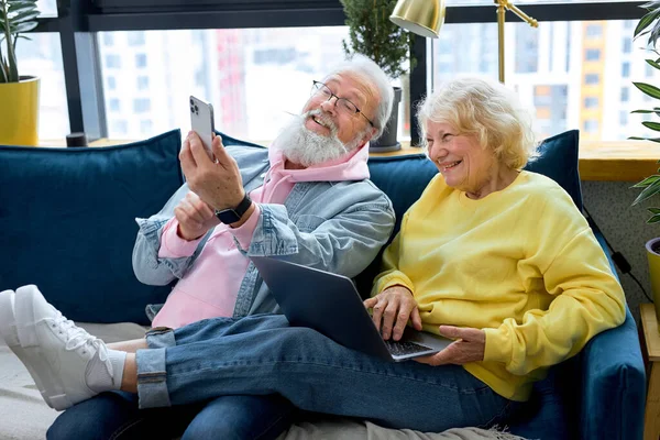 Happy mature retired couple using laptop and smartphone, reading news or watching tv series online. aged caucasian people have fun together at home, old people and modern tech concept