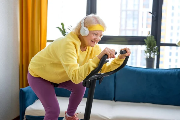 elderly caucasian woman training and doing exercise on cycle at home. active pensioner lifestyle concept. Retired woman training with stationary bicycle, doing workout and physical exercise