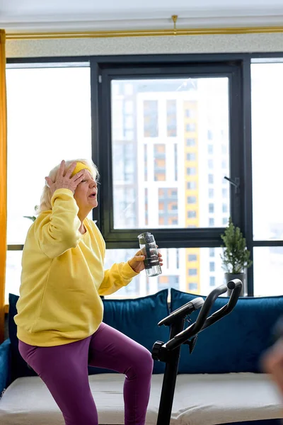 Elderly person training on stationary bicycle doing physical exercise and activity. aged caucasian woman using cardio cycling machine to train legs muscles with gymnastics at home, tired