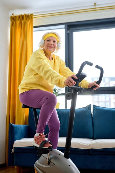 happy elderly person training on stationary bicycle doing physical exercise and activity. Senior caucasian woman using cardio cycling machine to train legs muscles with gymnastics at home.