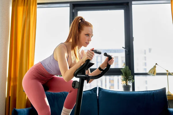 confident redhead woman working out on exercise bike or stationary bike at home alone. Caucasian fitness woman in stylish sportswear. Sport, workout, cardio or healthy lifestyle concept.