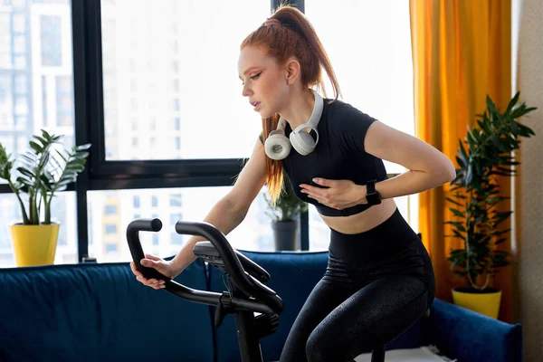 Home fitness workout woman training on stationary bike indoors. redhead woman working out at home on the exercise bike, hard to cycle, having pain, cycling for good health, engaged in sport