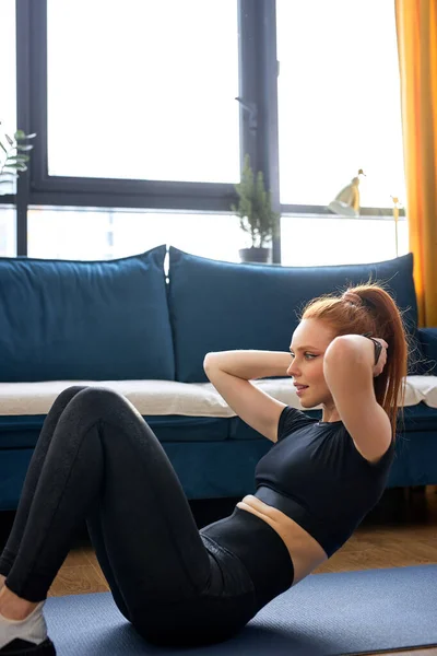 年轻迷人的 酷酷的女运动员穿着黑色运动服 在家里的地板上做健身锻炼 红头发的高加索女人 过着健康的生活 独自在室内锻炼 — 图库照片