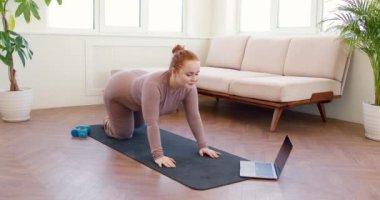 smiling plump woman raising legs, following online workout on laptop. Body Shaping And Glutes Workout. Woman In purple Sportswear Doing Donkey Kicks Exercise With Leg Raise Training Butt And Hamstring