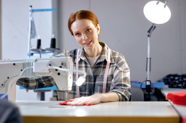 Modern endüstriyel dikiş makinesi kullanan, fabrikada çalışan beyaz kadın, atölyede çalışan, kameraya bakan kızıl saçlı hoş ve hatunlar. dressmaking, giyim üretim konsepti