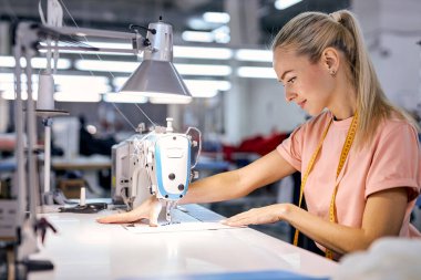 Modern dikiş makinesi kullanarak atölyede üretim hattında çalışan beyaz tenli bir tekstil fabrikası işçisi. Dikiş ya da terzilikle uğraşan çalışkan genç bir kadın, terzilik konsepti