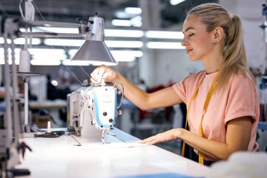 Genç bir kadın atölyede profesyonel bir makineyle dikiş dikiyor, giysi fabrikasında. Dikiş bölümünde kadın, terzi stüdyosu, dikiş okulu, kıyafet imalatı, dikiş endüstrisi.