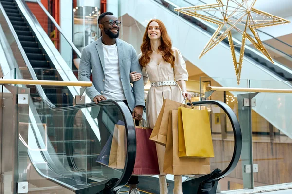 stock image young beautiful red-haired girl and african bearded guy get pleasure from buying goods, clothes, presents, gifts on holidays, escalator in the background of the photo