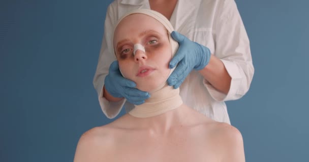 目にあざのある不幸な悲しい落ち込みの女の子は手術後です 隔離された青の背景 手術後に皮膚検査を実行する女性の顔に触れる遅い運動医師外科医 — ストック動画
