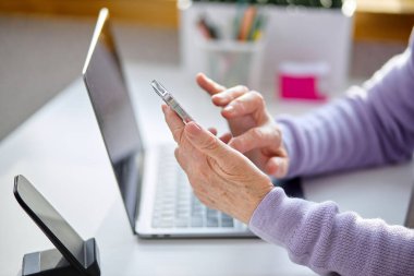 Akıllı telefon kullanan, akıllı telefon kullanan, e-posta haberlerini kontrol eden, oturma odasında oturan, internet ağlarında arkadaş arayan ya da bilgisayarda çalışan, blog yazan ya da Webinar izleyen yaşlı bir kadın.
