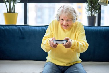 Sanal savaştan zevk alan, oyuna odaklanmış yaşlı bir kadın. Ev eğlencesi kavramı. Gündelik giyimli komik beyaz kadın evde kanepede oturmuş video oyunu oynuyor.