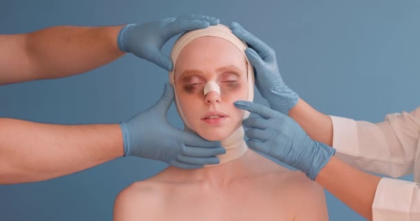医師の手は鼻 眼の下のあざの包帯でプレー落ち込んでいる女性の顔に触れる鼻形成術 閉鎖肖像分離青の背景 手術の医師の検査結果 — ストック動画