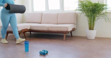 Elini kullanan tombul kızıl saçlı kadın yoga halısını açıyor. Bir şişe su ve dambıl yerde, spor için hazırlık, yoga yavaş çekim, yaşam tarzı, hobi