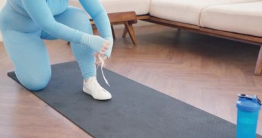 Mavi spor giyimli genç bir kadın ayakkabı bağcığı takıyor. Egzersiz yapan kadın antrenmana hazırlanıyor. Beyaz spor ayakkabılarını bağlıyor. Yoga minderinde oturuyor..