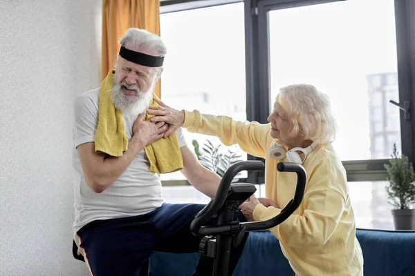 mature female helping man at home to do exercise on stationary bike. active couple of pensioners in sportswear help each other, male having pain, hard to do workout on sport equipment
