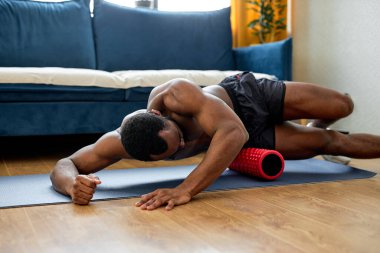 Yakışıklı, kaslı ve üstsüz siyah adam spor malzemeleriyle yerde antrenman yapıyor, evde tek başına. Koyu tenli erkek spor yapıyor, sağlıklı bir hayat sürüyor, formda ve güçlü bir vücut yapısı var.