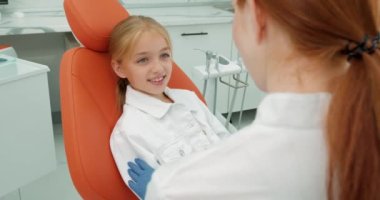 Mutlu cesur beyaz çocuk diş sağlığı kliniğinde diş hekimi, yan görüş ve tedavi öncesi beyaz üniformalı profesyonel stomatolog dişçiye bakarken ağzı açık bir şekilde sandalyede oturuyor.