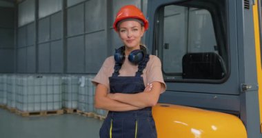 Fabrika lojistik nakliye deposunda forklift şoförünün yanında gülümseyen kadın işçi. Başarılı bir iş çıkardıktan sonra kolları bağlı, üniforma giymiş ve turuncu koruyucu şapka takmış bir şekilde poz veriyor.