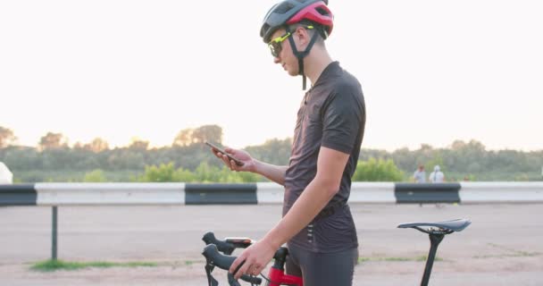 穿着运动服 戴头盔 戴眼镜站在路上 用手机发短信 用在线应用程序搜索Gps坐标的快乐英俊的男性骑自行车者旅行 运动现代技术 — 图库视频影像