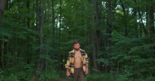 留着胡子 头戴斧头的英俊疲倦男子在绿林中散步 呆在树林里 看着相机 微笑着慢动作 寻找倒下的树生火的家伙 — 图库视频影像
