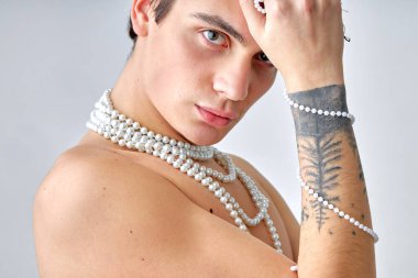 Kafkas erkek makyajı. Beyaz arka planda kameraya bakan üstsüz genç adamın yakın plan portresi. Elinde kolyesi ve dövmesi olan yakışıklı bir gay. insanlar, moda, güzellik konsepti