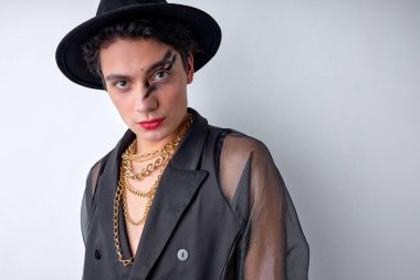 Erkek makyajı. Makyajlı, şapka ve zincirli, kameraya bakarak poz veren modern havalı adamın portresi, şık bir adam efemine görünüyor. Beyaz stüdyo arka planında izole edilmiş.