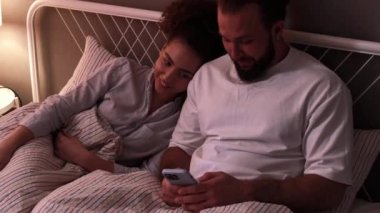 Mutlu çift yatakta uzanmış akıllı telefon kullanıyor, internette birlikte sörf yapıyor, eğleniyor, neşeli erkek, kadın sohbeti, sohbet, yatak odasında cep telefonu kullanıyor. Çift, evdeki cep telefonuna bakarak konuşuyor.
