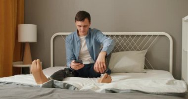 Protez biyonik bacakları olan neşeli mutlu genç adam cep telefonu kullanarak internette geziniyor online alışveriş yapıyor ve yatakta oturuyor. Boş zaman yaşam tarzı Yavaş çekim