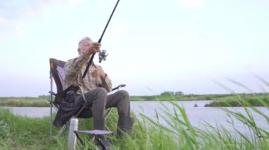 Yaşlı ciddi balıkçı oltayı tutuyor ve oltayı çeviriyor, ağır çekimde. Adam balık yakalıyor, gölde balık tutarken olta çekiyor. Vahşi doğa. Rüzgarlı yaz günü Tatil