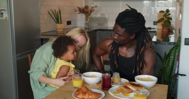 可爱可爱的非洲小女儿坐在妈妈的膝上慢动作妈妈给女儿吃果汁和食物 在舒适的厨房里 漂亮的黑人孩子在家里吃早餐时喝着新鲜的果汁 — 图库视频影像