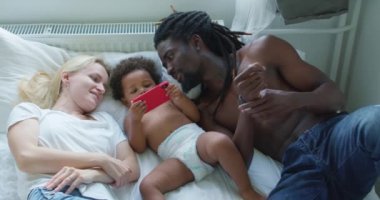 Küçük sevimli siyah çocuk çizgi film seyrediyor cep telefonuyla video çekiyor evde yatakta yatıyor, ebeveyn kızı oturma odasında dinleniyor, cihaz ekranına bakıyor yavaş çekim yaşam tarzı beleş