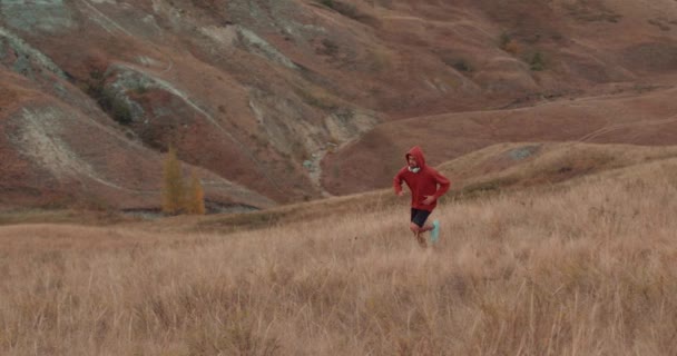 秋天里 田径运动员在高山上奔跑 动作缓慢 戴着红色帽衫头戴耳机的男人 日出时穿着毛衣跑上山 傍晚时分落日 — 图库视频影像