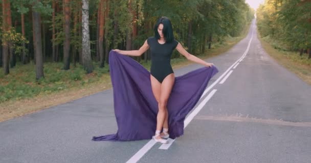 黒のボディスーツを着た若い女性のバレリーナは 紫色の布で点で踊り 足を上げ 手で布を振って 道路上の森の中で踊ります スレンダーな女性は完璧な体を持ちバレエダンスだけを — ストック動画