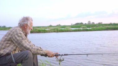 Çek tişörtlü yaşlı sakallı adam nehir kıyısında oturur ve balık tutar. Kıdemli balıkçı nehirde balık yakalar.