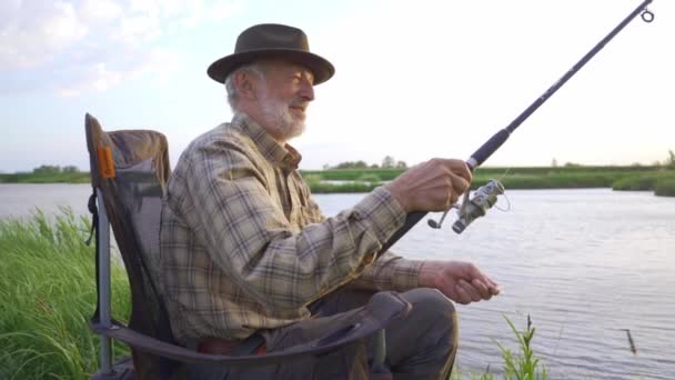 美しい湖で釣りを楽しむ帽子の古い祖父 屋外の男は釣りのプロセスから喜びを得ます ライフスタイル無料の余暇趣味の利益スローモーション — ストック動画