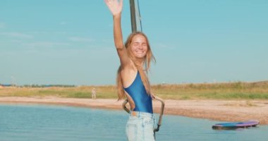 Neşeli kız rüzgar sörfü öğreniyor gözlerini kısıyor, merhaba diyor, selam veriyor arkadaşlara, seyahatten zevk alıyor yaz tatili ağır çekim jestleri yapıyor.