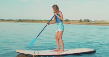 Gölün arka planındaki muhteşem doğa manzarasında Paddle Sörf Tahtası 'nda yoga yapan güzel bir sporcu kadın eğitimi. Fıstık gibi kız kürek çekiyor. hobi yaşam tarzı boş zaman