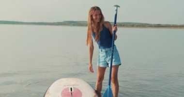 Mutlu, güler yüzlü, mayo giyen ve elinde kürekle şort giyen bir kadın SUP sörfünden sonra dinleniyor. Gün batımında yavaş çekimde tatilin tadını çıkarıyor.