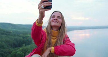 Gülümseyen sarışın yürüyüşçü kayanın üzerinde otururken akıllı telefondan selfie çekiyor. Neşeli kadın tatil gezisinde açık hava yaşam tarzını seviyor. Uçurumun kenarında oturan akıllı telefonla denizleri fotoğraflayan bir kadın.