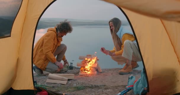 観光客 火災で暖かいカップルの手を愛します彼らは火の周りに座っています 旅行のコンセプトファミリーは新しい場所 キャンプを楽しむ 川を背景に 前景のテントで夜に木を燃やす — ストック動画