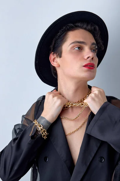 Attractive young transgender man LGBTQ being as woman posing with chain, in hat, gorgeous caucasian guy showing self-confidence, with red lipstick and make-up isolated on white background.