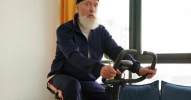 Gri saçlı yakışıklı yaşlı adam evde egzersiz bisikleti eğitimi alıyor, sağlığını düşünüyor, yaşlılığa ayak uyduruyor. Yaşlı adam iyi durumda. Sert ve hızlı bir bisiklete biniyor. Ağır çekim.