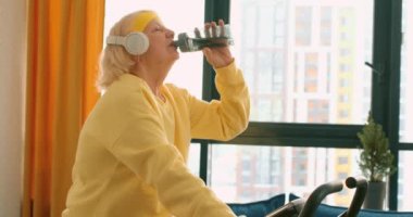 Yaşlı, terli, susamış, kulaklıklı bir kadın evde bisiklet sürüyor, su içiyor, şişe tutuyor, antrenman sırasında dinleniyor ağır çekimde dinleniyor, boş zaman hobileri var.