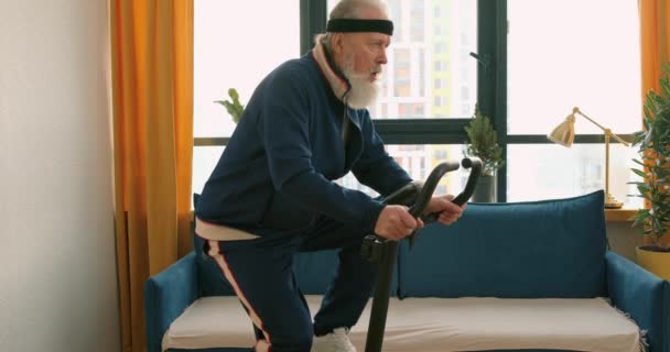 身体運動活動を行う固定自転車の老人ベアードマントレーニング自宅で体操で脚の筋肉を訓練するためにカーディオサイクリングマシンを使用して成熟した男 — ストック動画