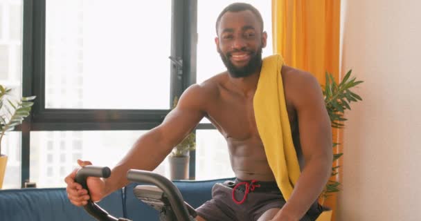Uśmiechnięty Aktywny Przystojny Afroamerykanin Sportowiec Odpoczywa Siedząc Stacjonarnym Rowerze Domu — Wideo stockowe