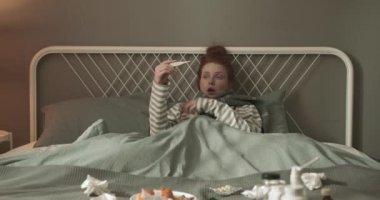 Zavallı, genç, güzel, kızıl saçlı bir kadın. Bir yatakta virüs hastalığı ve ateşi var. Coronavirus covid-19 salgını konsepti. Depresyondaki kız yatakta otururken termometre tutuyor.