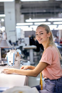 Genç beyaz tenli, sarışın tekstil fabrikası işçisi atölyedeki modern dikiş makinesini kullanarak üretim hattında. Çalışkan kadın dikiş ya da terzilikle meşgul, kameraya bakıp gülümsüyor.